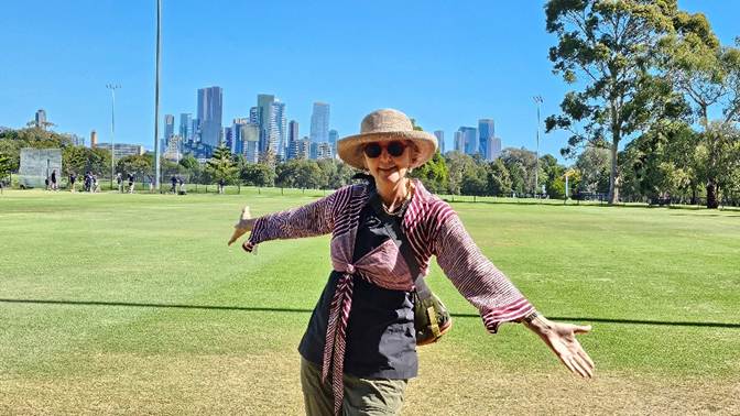 A person in a hat and sunglasses standing in a park with a city in the background

Description automatically generated